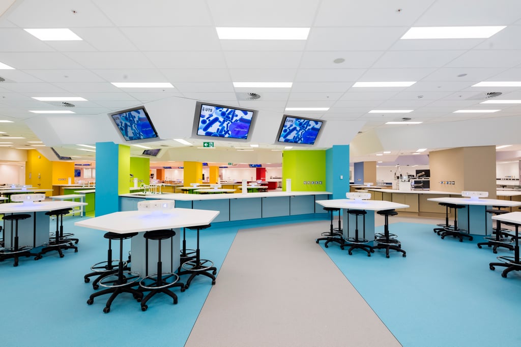 Interior view of the Superlab. Blue coloured floor with five hexagonal tables with black high stools on wheels interspersed around the area. Several large screens hung on the ceiling overlooking a large working desk. The working desk is connected to large lime and blue coloured columns.