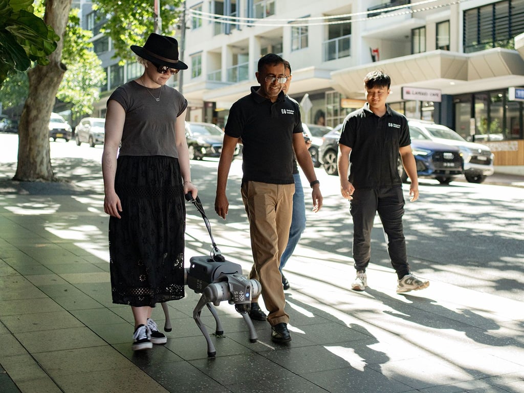 Zara Perry walks on a street in Sydney with assistance from the robotic guide. Sarath Kodagoda and a member of his team are present alongside her.