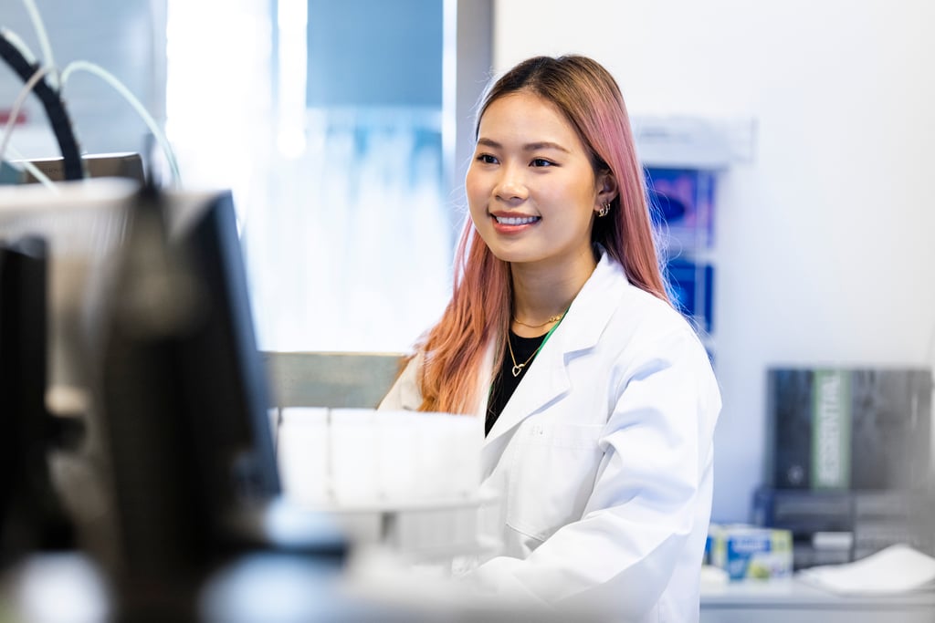 A female student with pink hair, wearing a lab coat and smiling at her desktop computer.