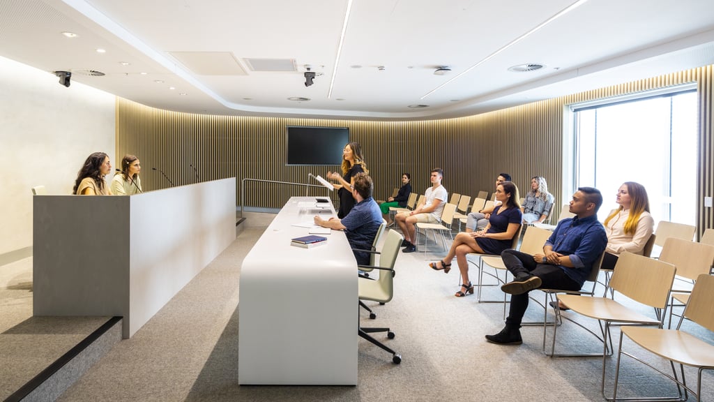 In a court setting. Two judges are focussed on the lawyer who is speaking. The client and the audience are watching the lawyer.