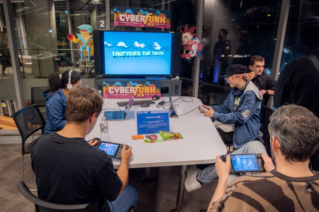 Students testing a Tech Fest video game project. Students are seated around a table with a digital screen playing on video games devices