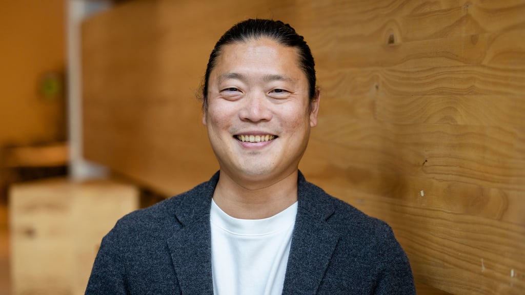 TaeWoo Kim stands centre frame smiling to the viewer. He is wearing a black blazer with white shirt. Behind him is the wooden blocks of the UTS Business School Oval classroom.
