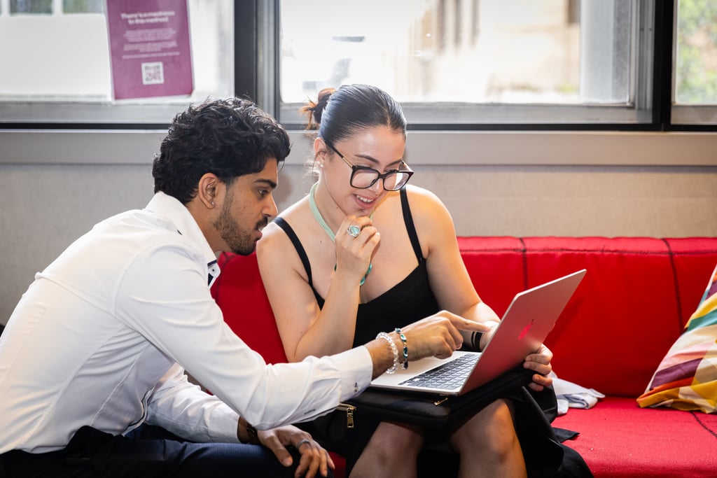 A man and woman sit on a red couch, engaging with a laptop. The woman, in a black dress, smiles and points at the screen.