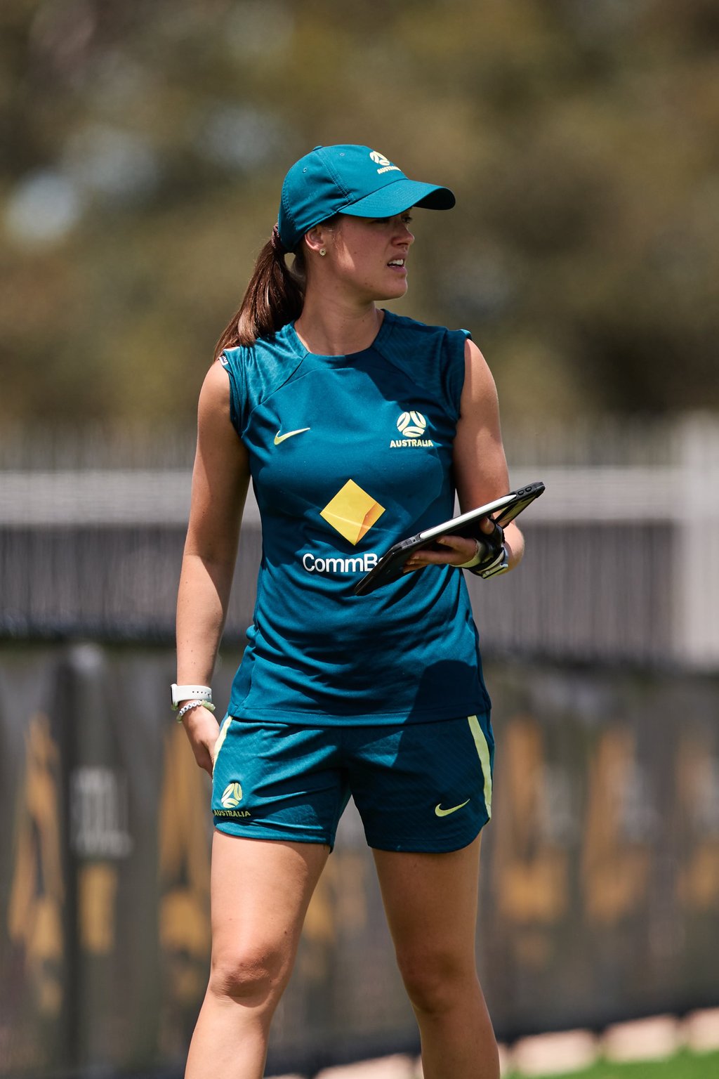 A woman in a green uniform and cap holds a clipboard, looking out at the field