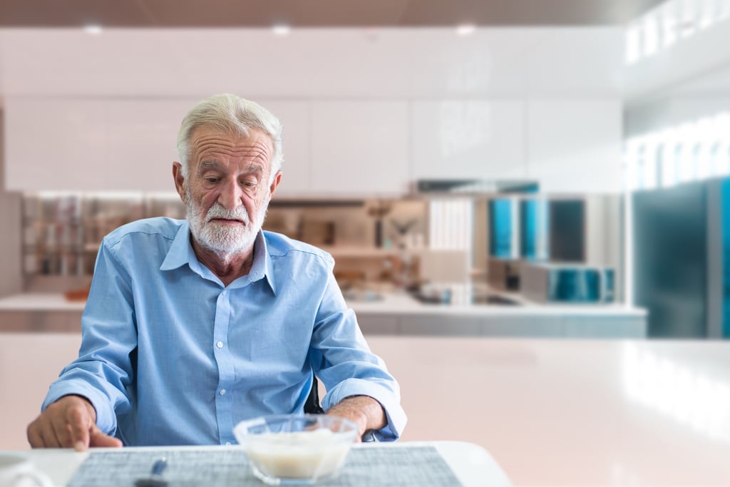 Elderly man sitting quietly at a kitchen table, looking uninterested in his meal
