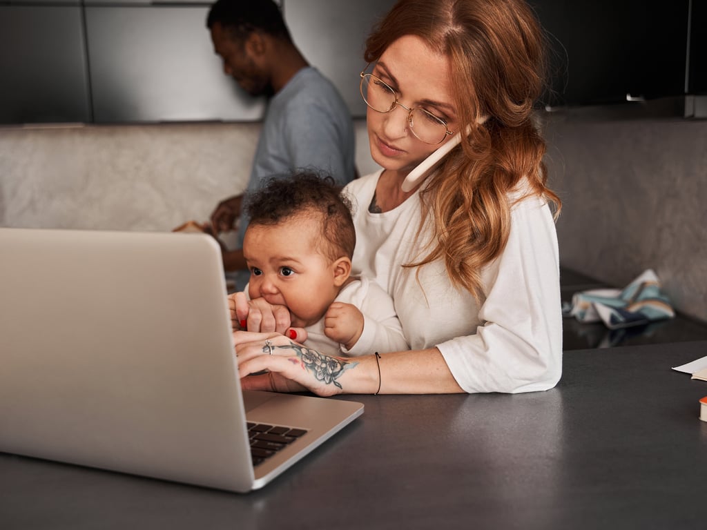 A mum with her baby on her lap taking a work call on her mobile phone, while her husband is cooking in the background.