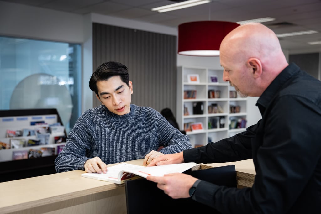Two men are engaged in discussion over an open book at a library counter. The space is modern and well-lit, conveying focus and collaboration.