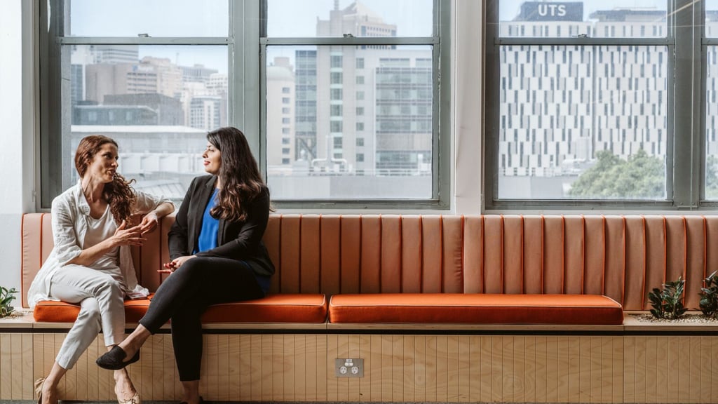 Two people sitting in front of windows making conversation, with UTS buildings in the background.