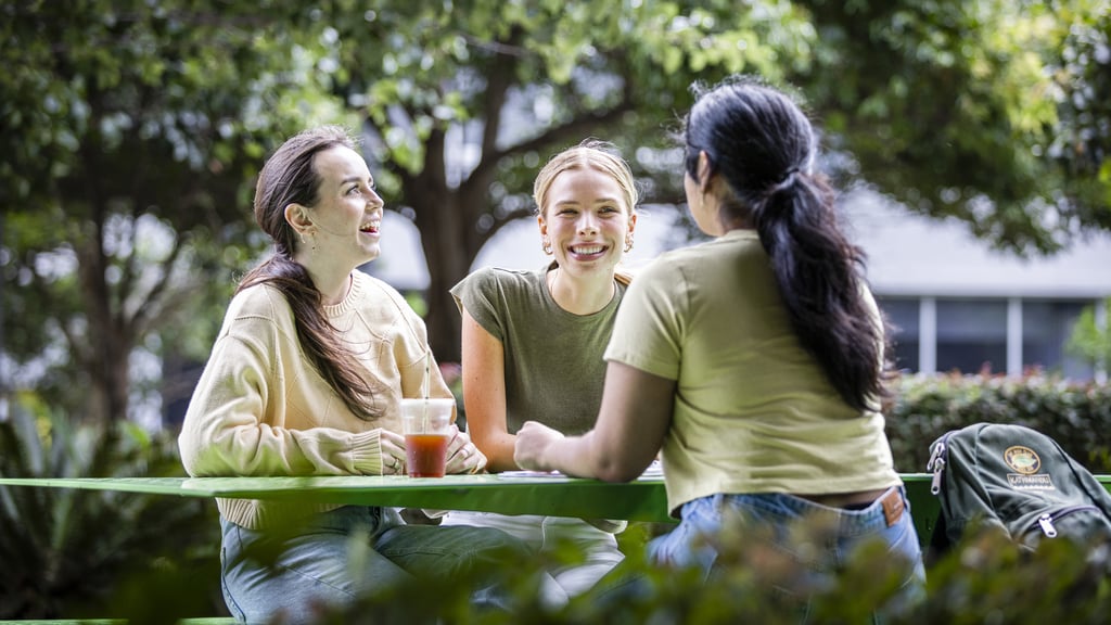 Three undergraduate female students talk on tables near Alumni Green, smiling and casual