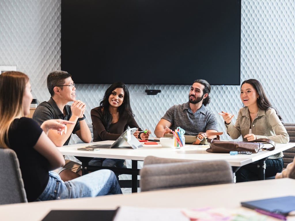 Four female and two male students sitting around a table discussing ideas. Laptop and other stationery on table.