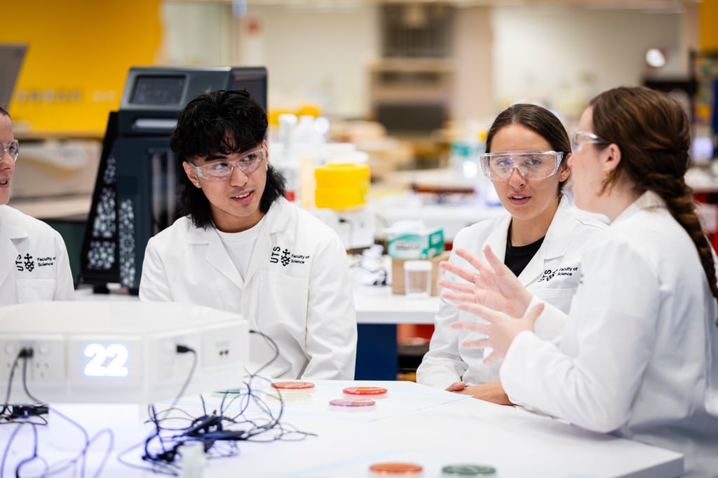 In the UTS HIVE Super Lab, four people in lab coats and safety goggles engaged in discussion at a lab table with wires and petri dishes.