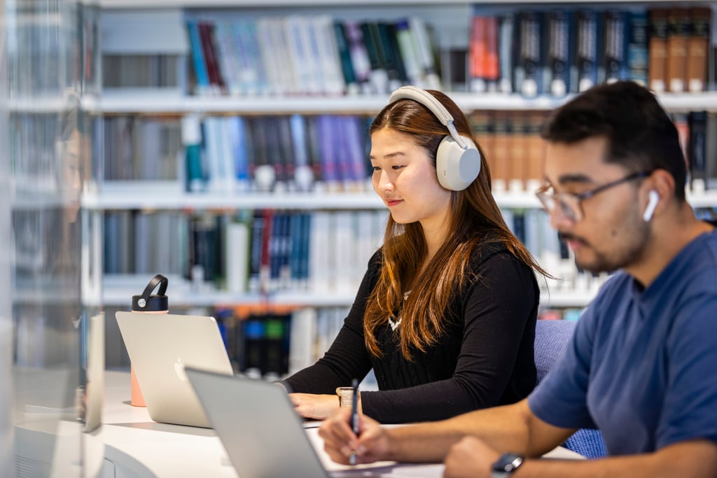 Two students focus on laptops in the UTS library. A woman wearing headphones is in the foreground, with bookshelves behind them, creating a studious atmosphere.