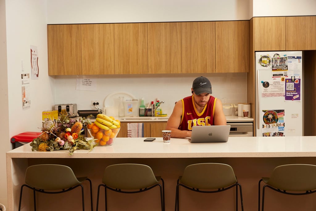 A man seated at a kitchen counter, focused on his laptop, surrounded by a cozy kitchen environment.