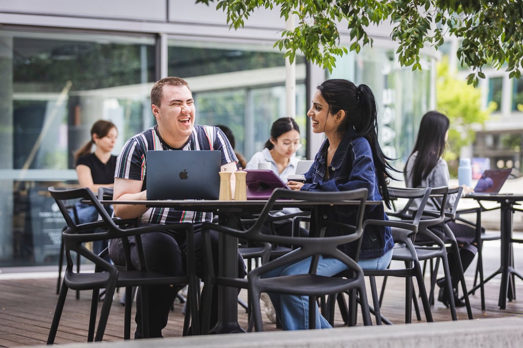 Two students sitting at a cafe with laptops in front of them, talking and laughing.