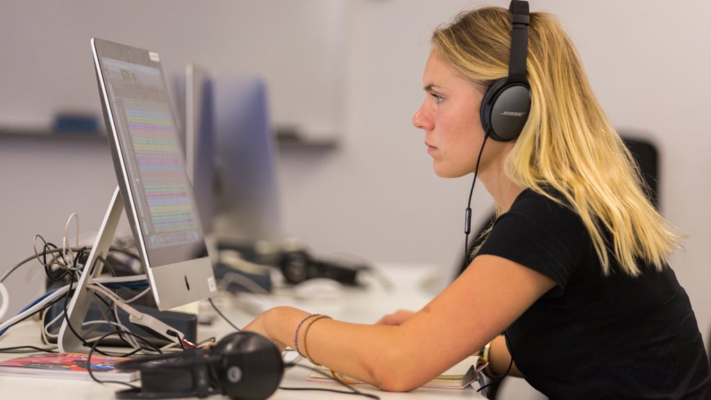 A woman with headphones intensely focuses on a computer screen displaying colourful audio waveforms. She is in a bright room with a calm, professional atmosphere.