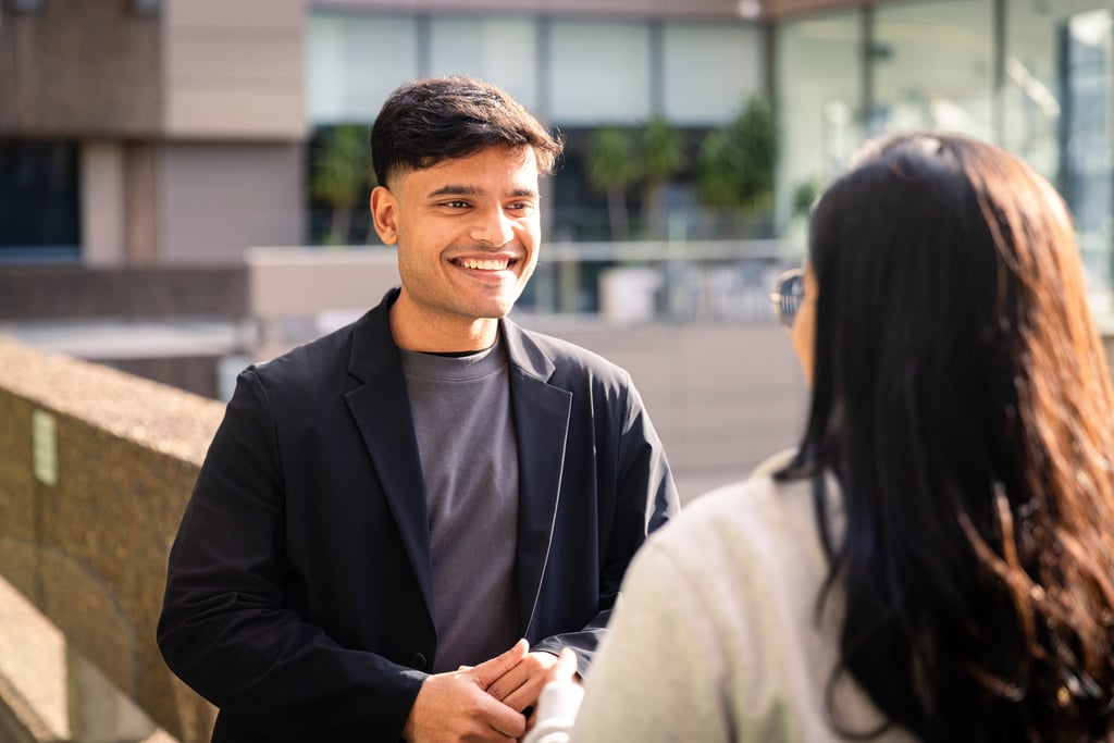 A male student in a black blazer smiles warmly while talking to another student. They are outside UTS Tower balcony on a sunny day, creating a friendly atmosphere.