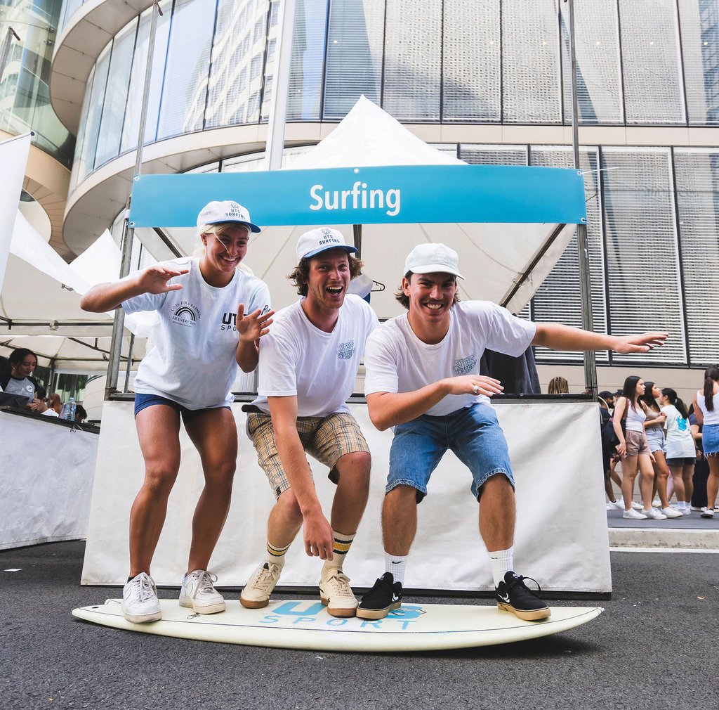 Three students on campus riding a surfboard in front of an event stall