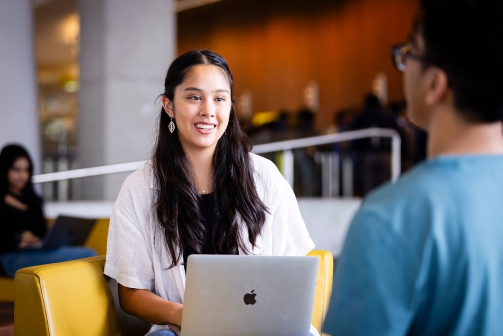 A young woman with long dark hair smiles while working on a laptop, sitting in a bright, casual setting. She engages with a person in a blue shirt.