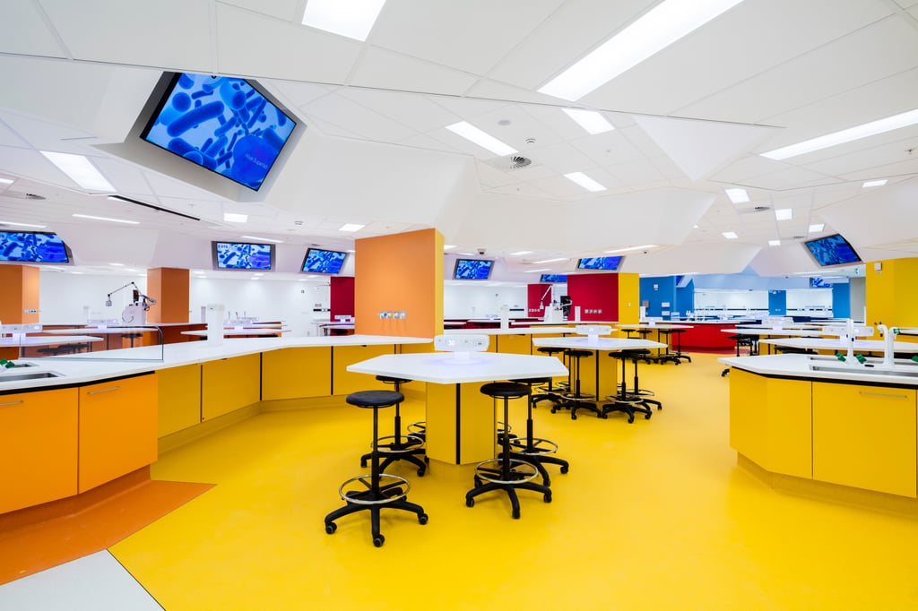 Interior of the Superlab. The floor is bright yellow, large working desks' bottom panels and columns are bright yellow and orange. There's large screens hung from white ceiling. Hexagonal shaped desks with black high stools on wheels.