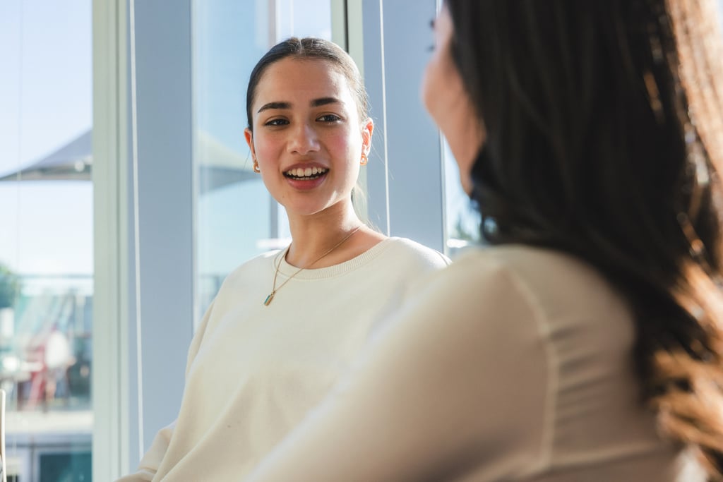 A woman in a white sweater smiles while talking to another person in the UTS Library, with bright sunlight streaming in through large windows behind them.