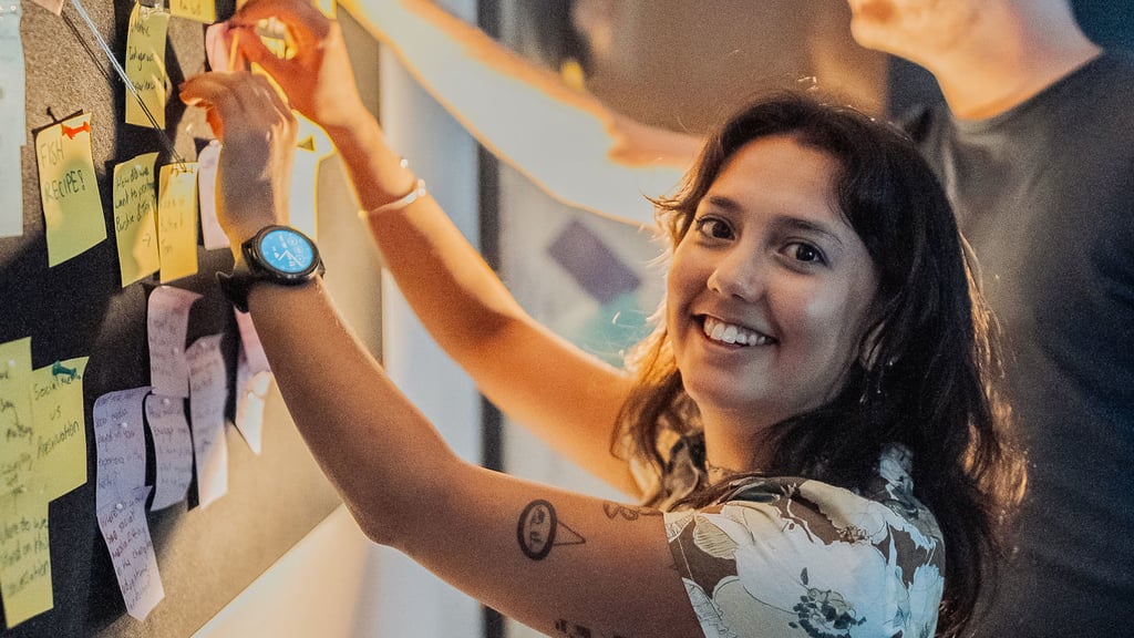 A smiling UTS student holds a sticky note against a wall, reflecting enthusiasm and creativity in her workspace.