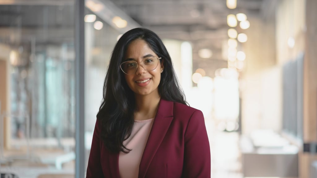 In the foreground Vani, a young woman wearing glasses and a burgundy blazer, is smiling confidently at the viewer. In the background is a bright, modern office hallway at UTS Business School.