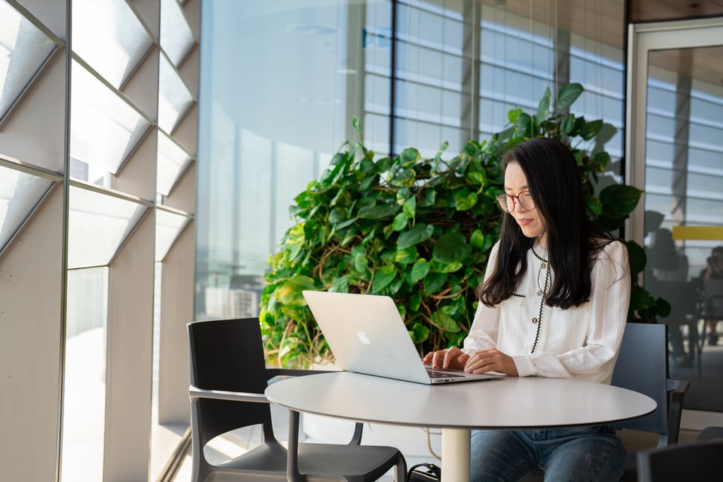 Female international student studying quietly on her laptop in front of a plant and glass windows.