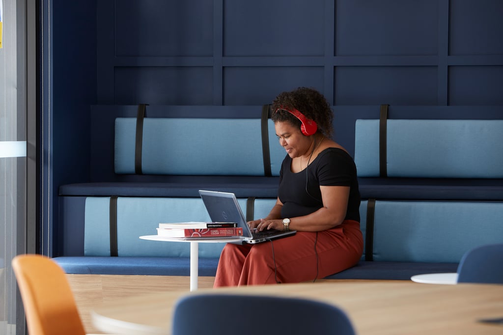 An indigneous woman wearing red headphones works on a laptop at a white table in a modern blue and gray seating area. She appears focused and relaxed.