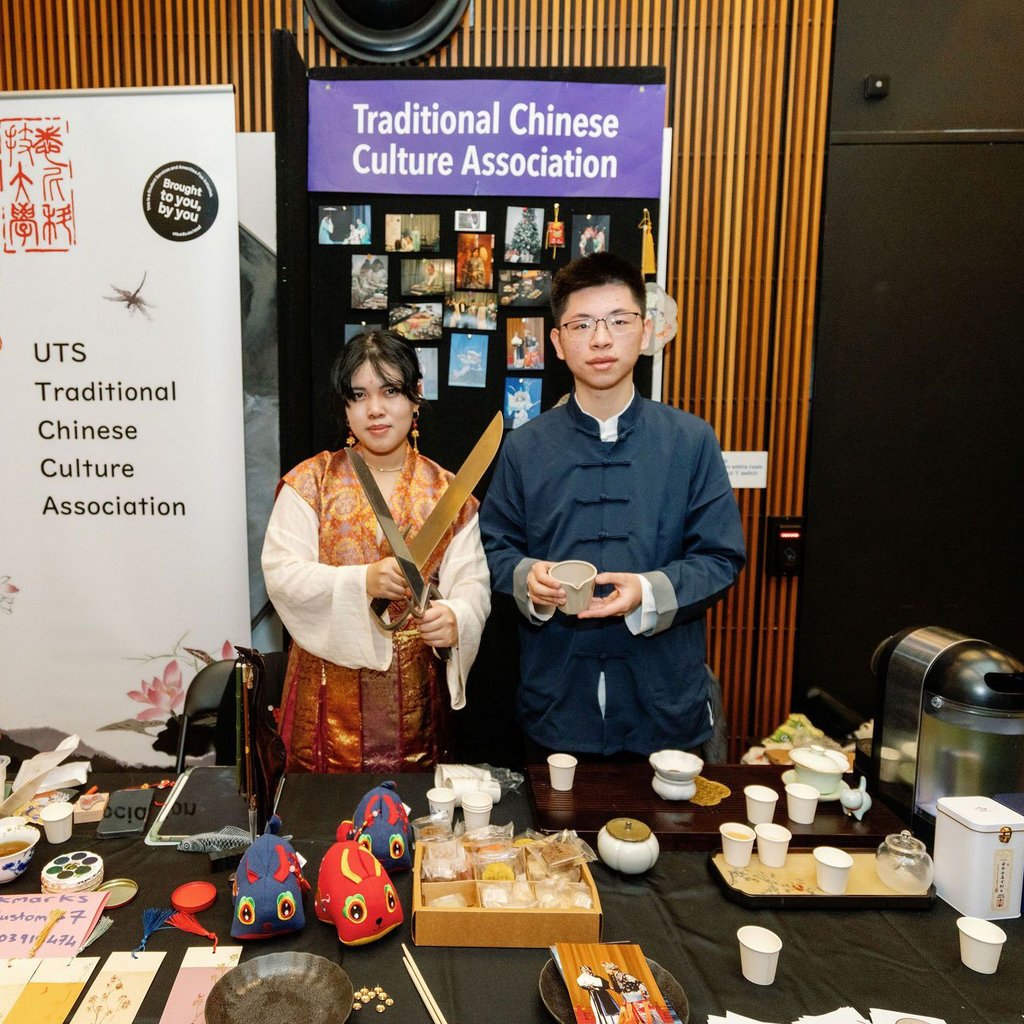 Two students standing at an event booth with the sign Traditional Chinese Cultural Association; one student holds two swords while the other holds a tea receptacle.