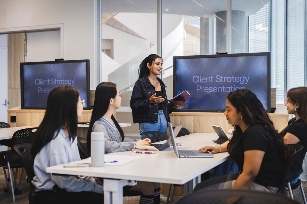 Group of students sitting in a class with large screens, one is standing and presenting.