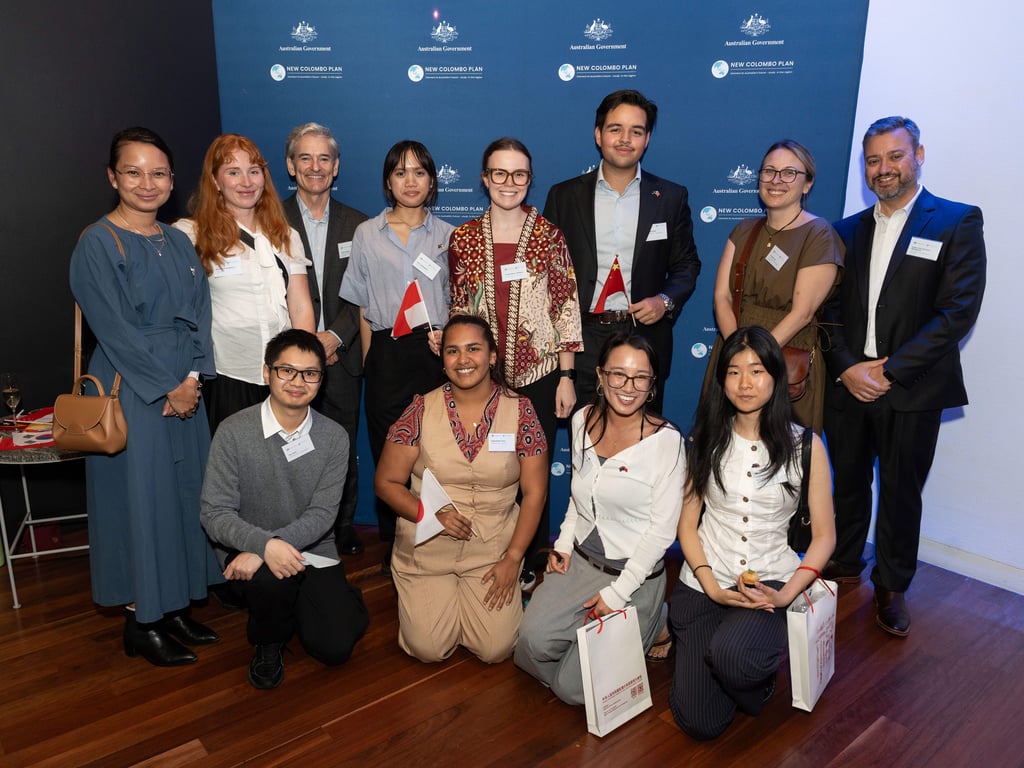 Group of scholars and representatives stand against blue media wall with Australian Government and New Colombo Plan logos. From left to right, top row: Kartika Paksi, Madeleine McWilliam, Innes Ireland, Nina Jayengrani, Amelia Wilson-Williams, Michael Farah, Alice Loda, Nick Manganas. Bottom row: Leo Chau, Kayla Baker-Peris, Emily Rutten, Lucia Guo.