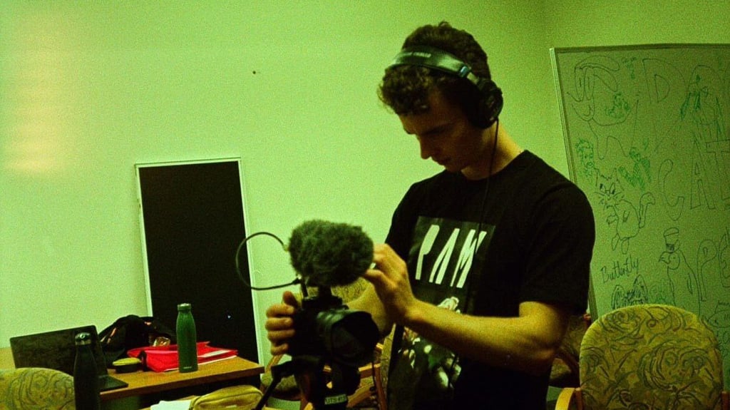 Harry Lewis standing in a green room wearing headphones and holding camera and microphone equipment.