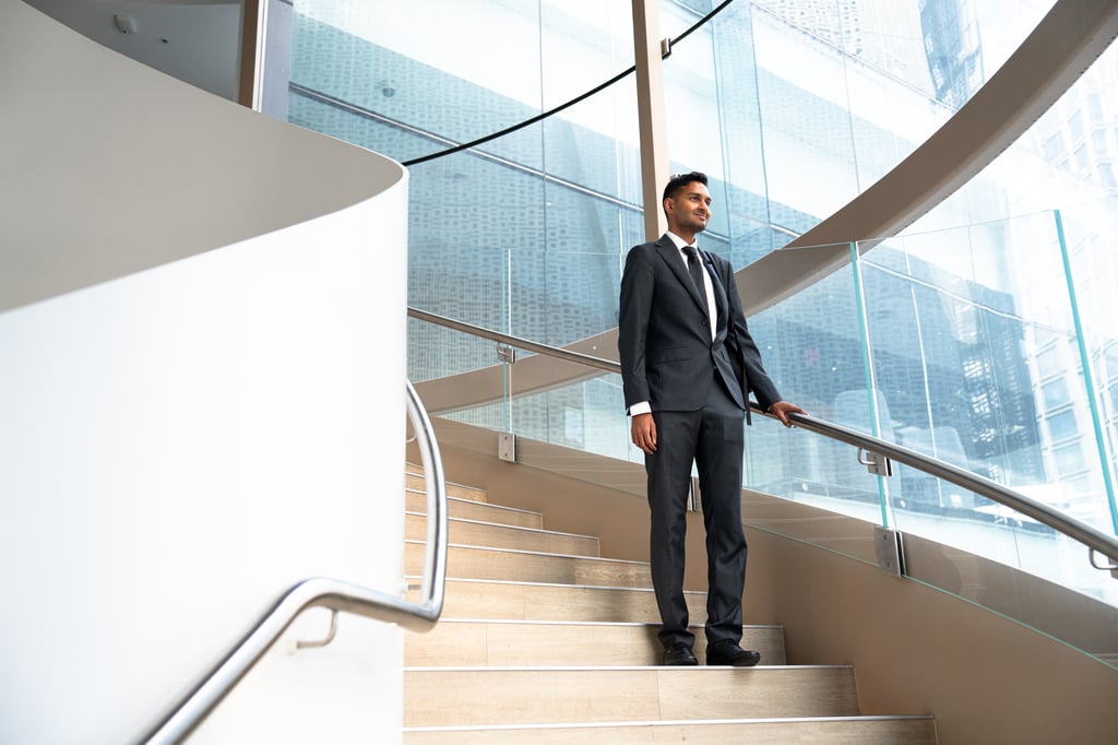 Sai wears a suit and tie, and stands on the spiral staircase