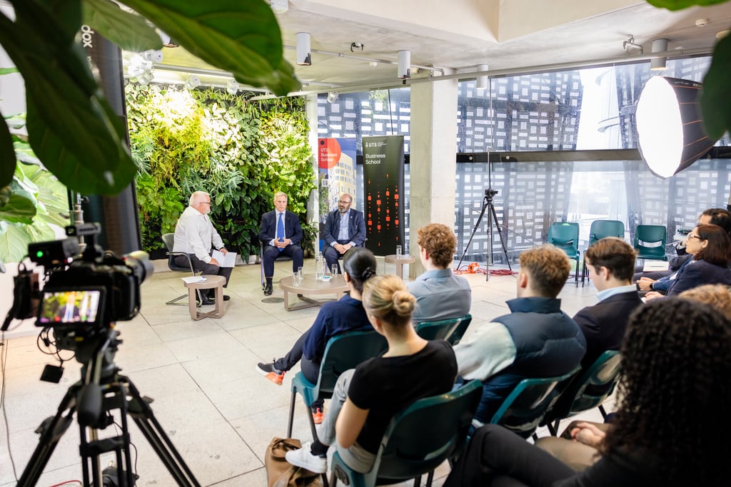 Foreground, a video camera and a collection of seated students sit with their backs to the viewer. In the background seated in front of the students are three speakers seated in chairs. One is dressed in a white shirt and black pants and the other two speakers are dressed in black suits. They are seated in a light filled room with lush plants growing off a back panel and glass windows overlooking the UTS campus.