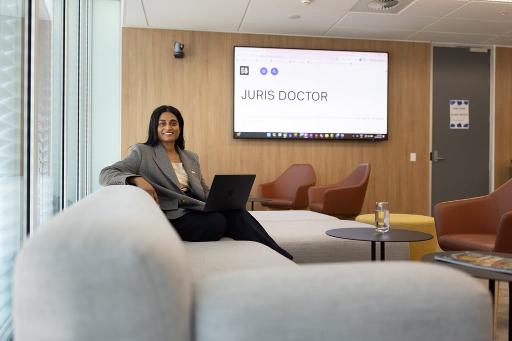 Ash, wearing a grey blazer, sits on a white couch using her laptop, in the postgraduate Law lounge