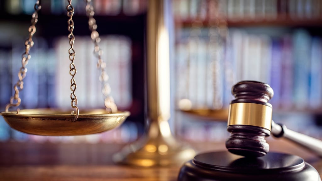 A wooden judge's gavel and brass scales of justice are on a desk in a library. The background of blurred books conveys a sense of authority and law.