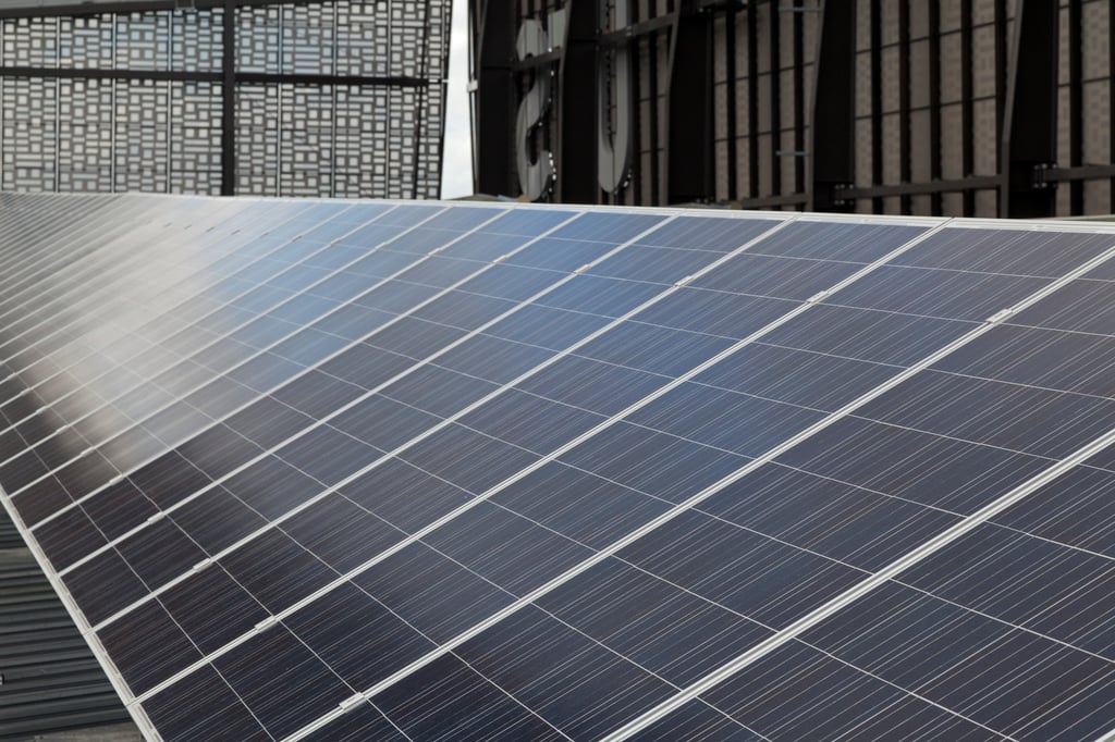 Close-up view of a large array of blue solar panels installed on a rooftop or industrial structure. The panels are arranged in neat rows and angled toward the light, with a modern building facade visible in the background, illustrating renewable solar energy generation.