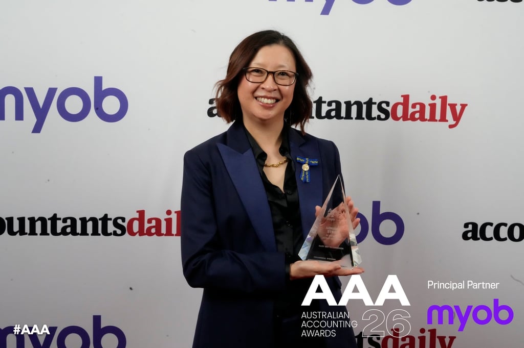 Amanda White stands in front of a media screen, smiling to camera while holding up her engraved award. She is wearing a blue suit with a gold necklace, gold ribbon pinned to her left breast pocket and glasses. The words "AAA Australian Accounting Awards 2026" appear beneath the image.