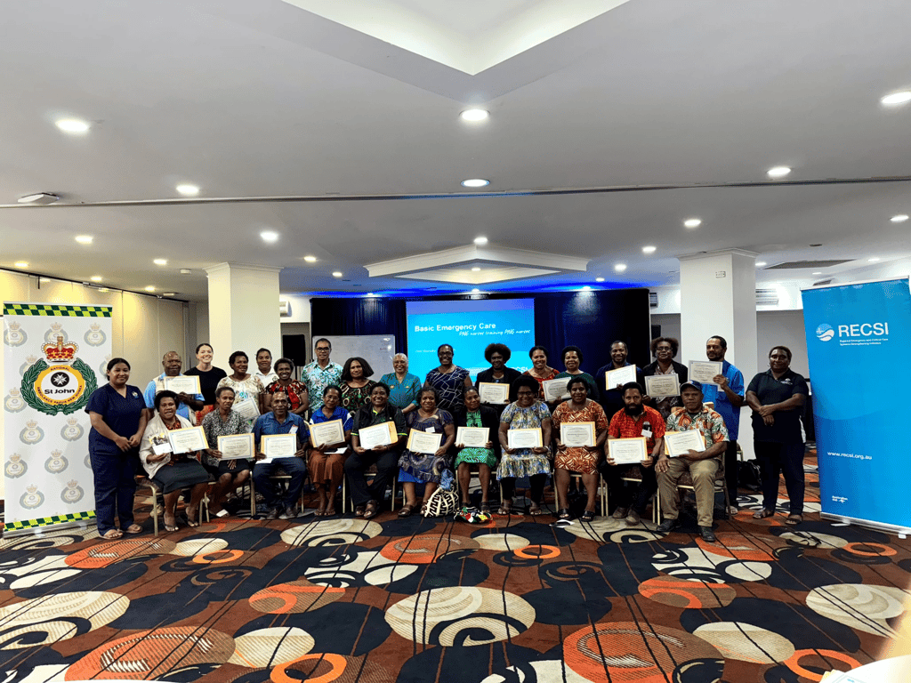 A group of 28 people, diverse in gender and attire, proudly display their certificates while posing in a well-lit room. Banners for RECSI and St. John are visible. Emotions of achievement and joy are conveyed.