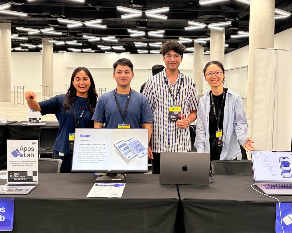 Four people stand behind a table with tech equipment at a startup summit. They are smiling and appear enthusiastic. The display shows a phone app interface.