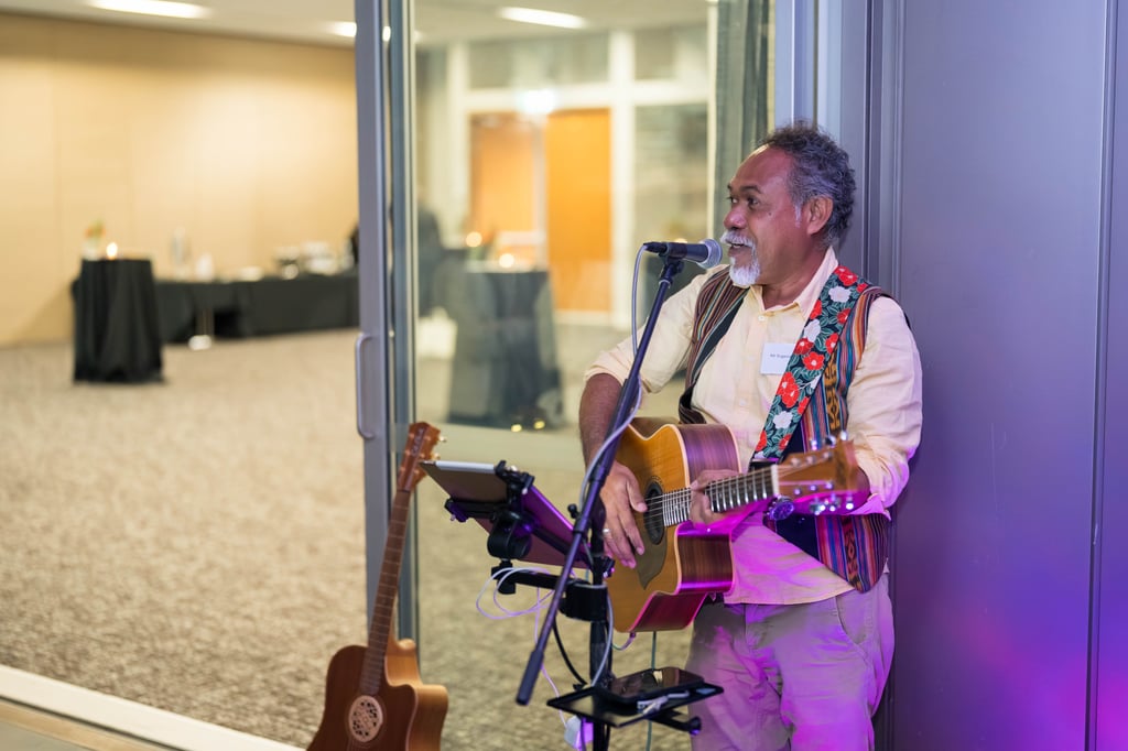 Musician with a patterned vest and colorful guitar strap sings and plays guitar near a glass door. The room is softly lit with purple tones, creating a relaxed atmosphere.