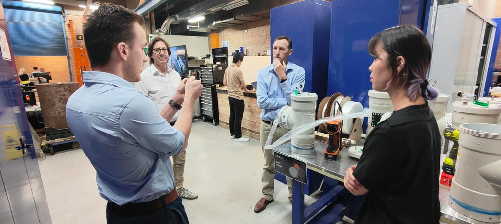 Four people in a workshop engage in discussion near a table with tools and equipment. The setting conveys a collaborative and focused atmosphere.