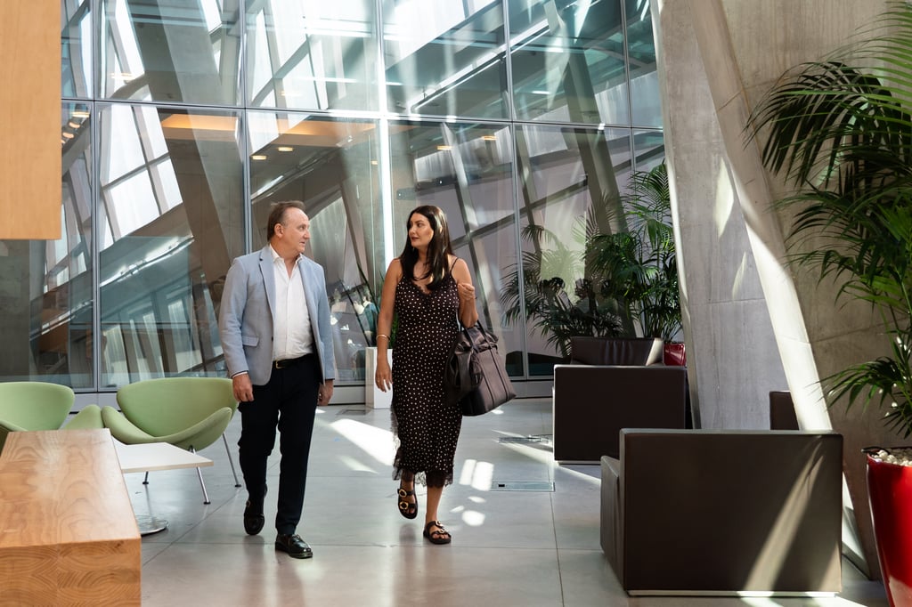 Alison Gibson walks and talks with Professor Andrew Heys through a light-filled modern building atrium with floor-to-ceiling glass windows and indoor plants.