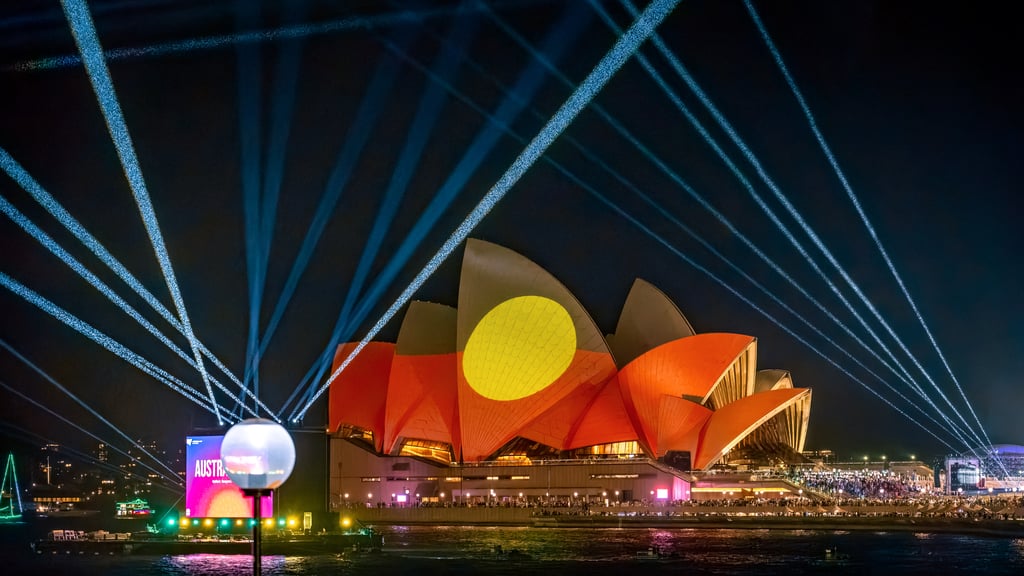 The Sydney Opera House is illuminated at night with vibrant red and yellow Indigenous flag art projections. Blue laser beams radiate into the night sky.