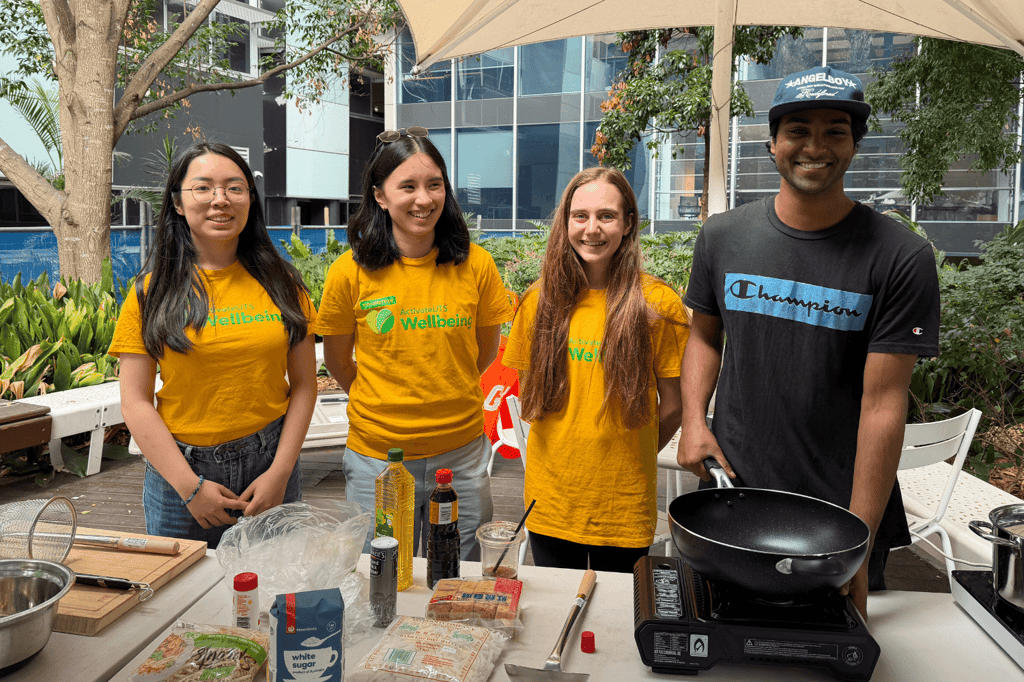 Four people standing behind an outdoor cooking station with a frying pan, cooking equipment and ingredients.