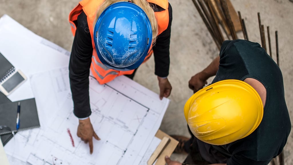 Overhead shot of two men wearing hard hats. The man with a blue hard hat is pointing to some blueprints spread on the table they are over while the man in the yellow hard hat watches on.