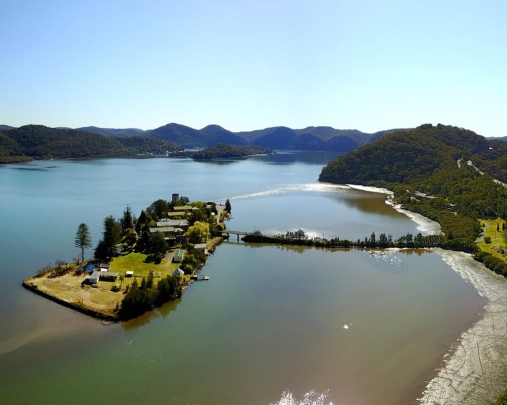 A scenic aerial view of a small, narrow island connected to the mainland by a bridge, surrounded by calm blue water and forested hills.