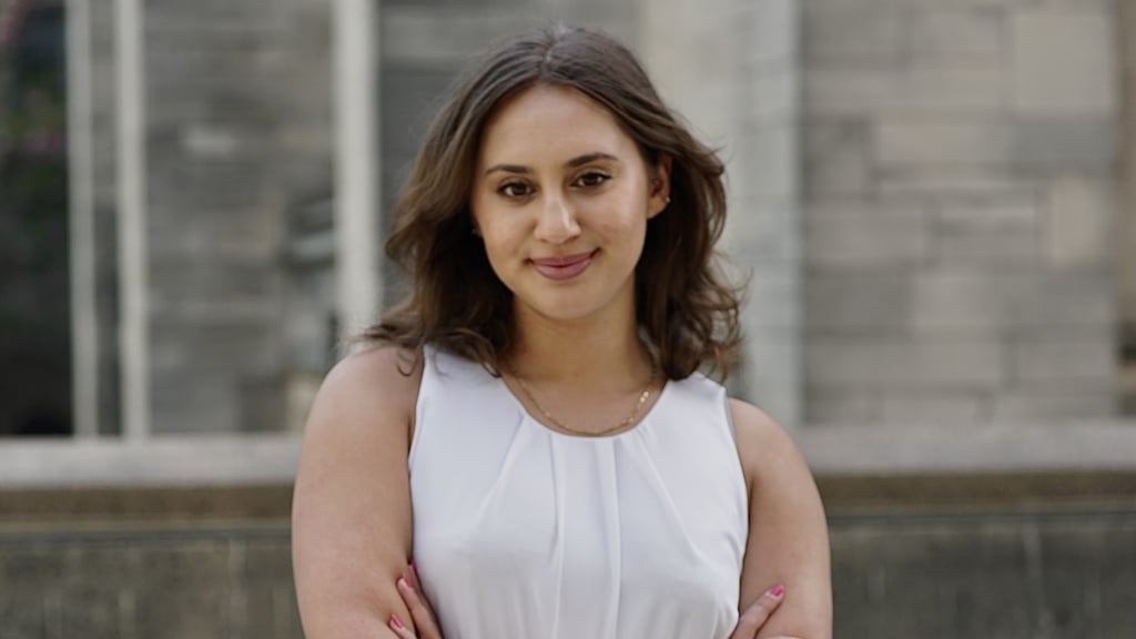 Jaya, wearing a white top and a gold necklace, stands in front of a historic stone building