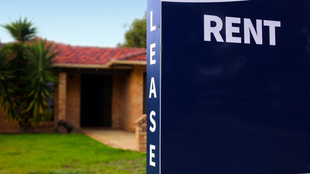 Close-up of a dark blue "For Rent" sign with "Lease" written vertically. A suburban house with a red tile roof and greenery is blurred in the background.