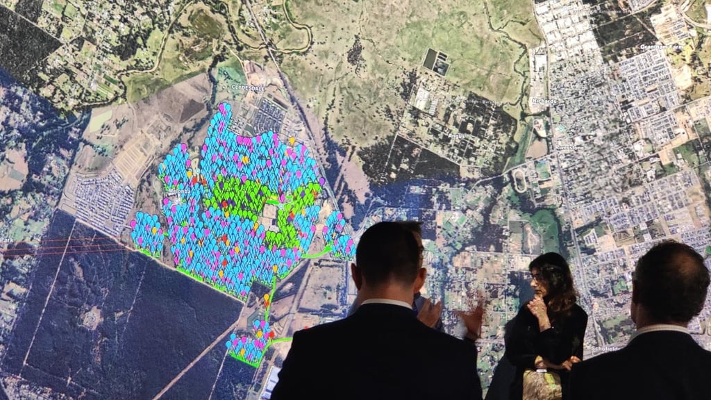 A small group of people stand in front of a large, detailed satellite map projection. The map shows a cityscape with colourful overlays, indicating data analysis.